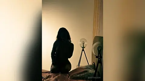 Silhouette of a woman in dark loungewear sitting on a bed in a moody, dimly lit bedroom setting.