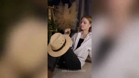 Young woman in a white shirt sitting by pampas grass decor, wearing a stylish beige sun hat in a cozy indoor setting.
