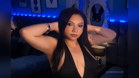 Young woman in a black halter dress posing in a moody bedroom with blue neon lighting.