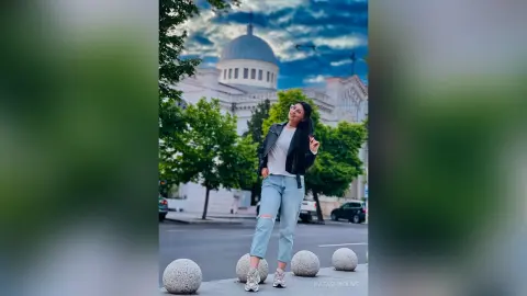 Stylish woman in a black leather jacket and light blue jeans posing in front of a grand white cathedral.
