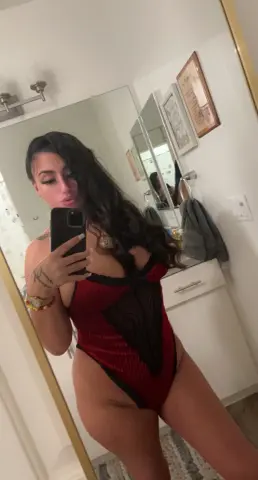 Woman in a red lace bodysuit taking a mirror selfie in a bright bathroom.