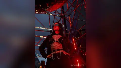 Edgy woman in black mesh outfit and sunglasses posing under red neon lights at a night event.