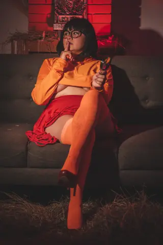 Woman in orange crop top and red skirt sitting on a couch in moody, warm lighting.