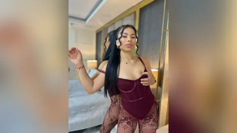 Glamorous woman in a burgundy lace bodysuit posing in a modern bedroom setting.