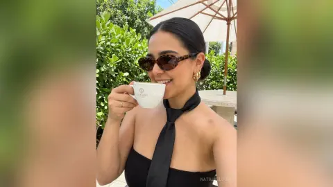 NatalyBloom smiles while wearing sunglasses and a black halter top, sipping coffee outdoors near green bushes.