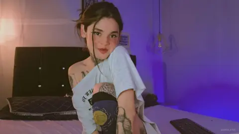 Young woman with tattoos in a white t-shirt relaxing in a bedroom with moody purple lighting.