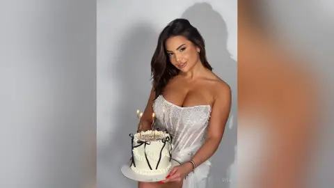 Smiling woman in a white sequin strapless dress holding a birthday cake with lit candles against a plain background.