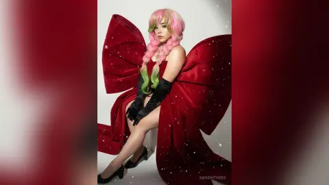 Cosplayer in a red velvet bow outfit and black gloves with pink hair posing against a white studio background.