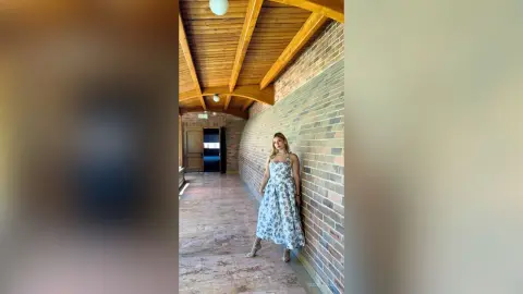 Woman in a blue floral maxi dress standing in a rustic brick hallway with wooden ceiling beams.
