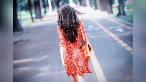 Young woman in a red polka dot dress walking down a sunny park path with a brown leather shoulder bag.