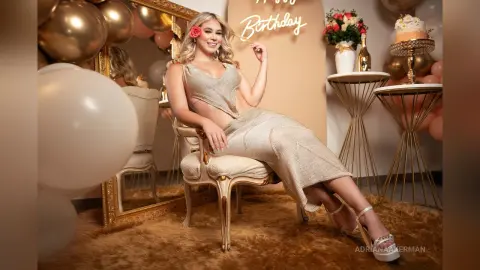 Smiling woman in a sparkling gold cocktail dress sitting in a glamorous birthday party setting with balloons.