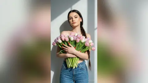 Beautiful young woman in denim jeans holding a large bouquet of pink tulips against a bright sunlit wall.