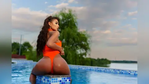 Anastasia poses in a bright orange bodysuit while sitting on the edge of a blue tiled swimming pool.