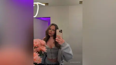 Young woman in a grey zip-up lounge set holding pink flowers in a modern bathroom with purple mood lighting.
