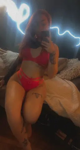 Red bikini set worn by a tattooed woman sitting on a bed in a moody, dimly lit room with neon light decor.