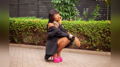 Stylish woman in a black outfit and pink platform heels posing outdoors near green garden bushes.