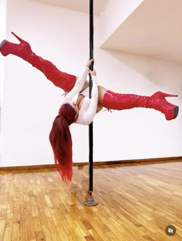 SabinaTriolo performs a pole dance move, wearing a white top and long red patent leather boots while hanging upside down.