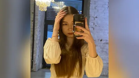 MagalyBoffa poses for a mirror selfie, wearing a long-sleeved cream blouse with her long brown hair styled down.