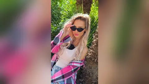 Stylish blonde woman in sunglasses and pink plaid shirt posing in a sunny tropical garden.