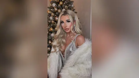 Glamorous blonde woman in a sparkling silver sequin dress and white faux fur coat posing by a Christmas tree.