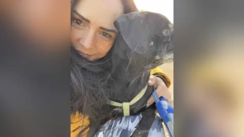 Smiling woman in a yellow jacket hugging a black dog in a warm, affectionate outdoor setting.
