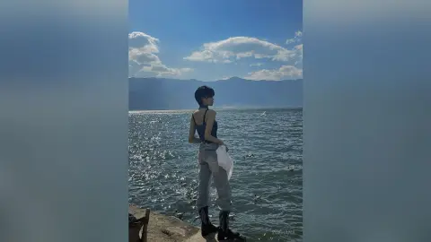 Young woman in a blue tank top and light jeans standing by a scenic mountain lake under a sunny sky.