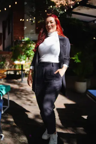 Confident woman in white turtleneck and black trousers standing in a sunlit outdoor cafe setting.