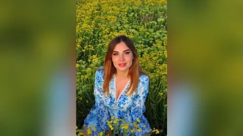 Smiling young woman in a blue floral dress sitting in a sunny field of yellow wildflowers.