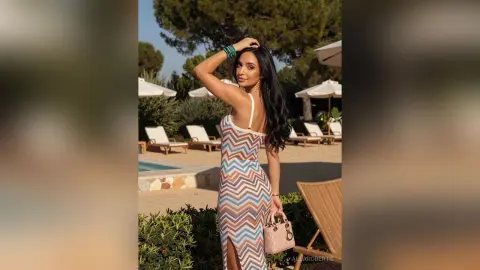 Woman in a patterned maxi dress holding a pink handbag by a sunny outdoor swimming pool.