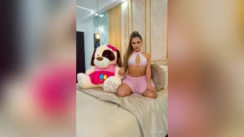 MiaBellini kneels on a bed wearing white lace lingerie and pink shorts next to a large stuffed dog.