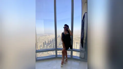 MissyJolie stands in front of a large window overlooking a city skyline, wearing a black tank top and black shorts with sunglasses.