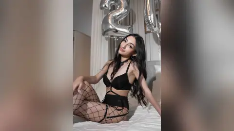 Young woman in black lingerie and fishnet tights sitting on a bed with silver birthday balloons.
