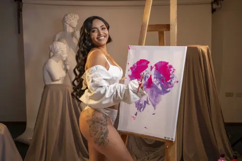 Smiling woman in white off-the-shoulder top painting a pink heart on canvas in an art studio with statues.