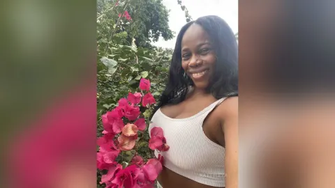 KattyLeya — KattyLeya smiles outdoors while wearing a white ribbed tank top next to bright pink flowers.
