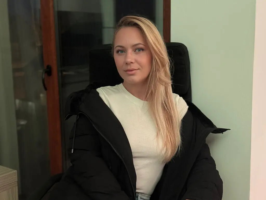 Blonde woman in a white t-shirt and black puffer jacket sitting indoors with a calm expression.