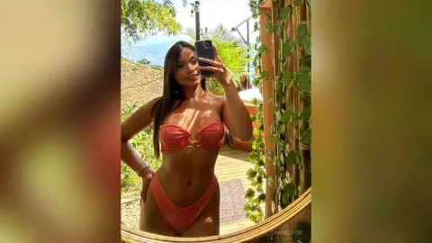 Smiling woman in a coral bikini taking a selfie on a sunny tropical balcony surrounded by lush green plants.