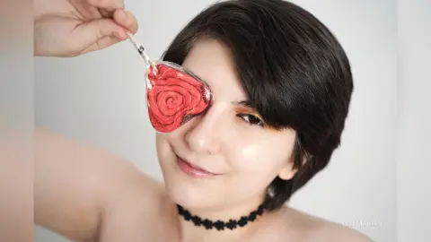 Young woman with short dark hair holding a red rose-shaped eye mask against her eye in a bright studio setting.