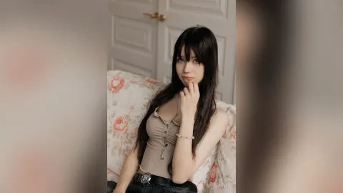 MintaTukes — MintaTukes sits on a floral bed with long dark hair wearing a tan ribbed tank top and black jeans.