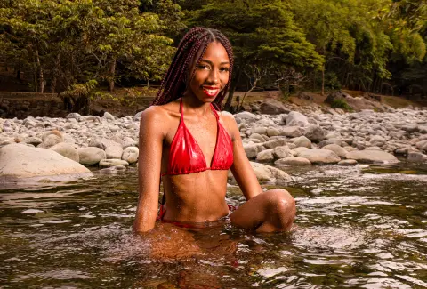 Smiling Black woman in a red bikini relaxing in a rocky river surrounded by lush green forest trees.