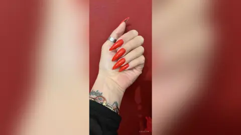 Close-up of a woman's hand with long red almond-shaped nails and silver rings against a red background.