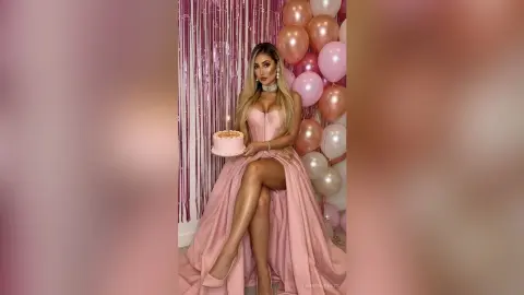 Glamorous woman in a pink evening gown holding a birthday cake amidst pink balloons and shimmering fringe curtains.