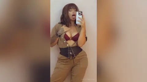 Curvy woman in a tan jumpsuit and burgundy bikini top posing playfully in a minimalist indoor setting.