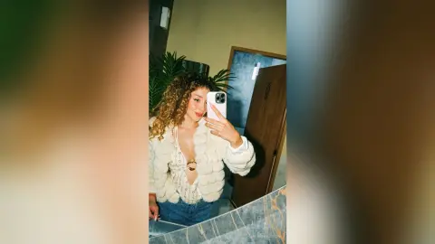 Curly-haired woman in a white fuzzy cardigan and denim jeans taking a mirror selfie in a cozy indoor setting.