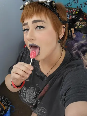 ChloeNightmare licks a pink lollipop with her tongue out, wearing a black t-shirt and short reddish-brown hair with bangs.