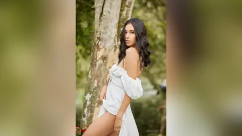 LucyMartin poses outdoors against a tree trunk wearing a white off-the-shoulder top that shows her bare back and shoulders.