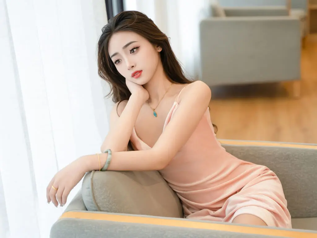 Young woman in a pink slip dress relaxing on a grey armchair by a bright window in a serene indoor setting.