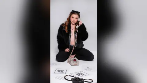 Young woman in a black faux fur coat holding a vintage telephone against a white studio background.
