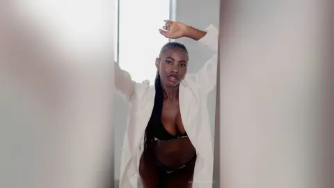 Stylish young woman in a white button-down shirt and black bikini posing confidently in a bright, airy room.