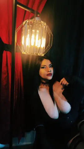 Young woman in a black outfit and choker posing under warm lantern light against red curtains.