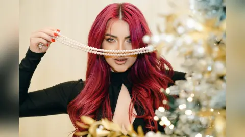 Glamorous woman with long red hair wearing a black dress and holding pearl necklaces near a festive silver tree.